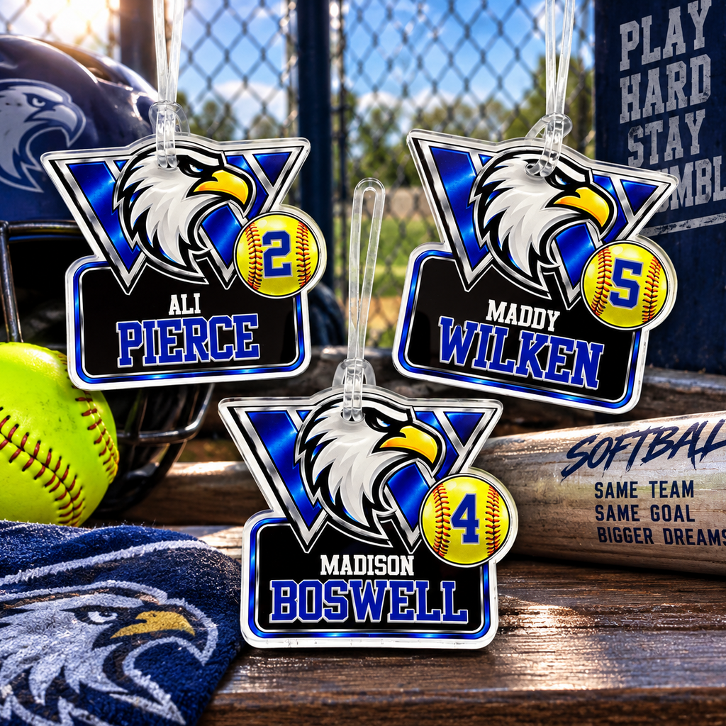 Three personalized acrylic hanging ornaments shaped like football helmets are displayed on a wooden surface outdoors near a chain-link fence. The ornaments feature eagle head graphics, blue and black backgrounds, player names (All Pierce, Maddy Wilken, Madison Boswell), and yellow baseball number circles.