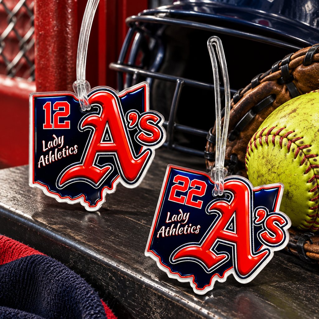 Two baseball-themed luggage tags featuring the '22 Lady Athletics A's' logo, displayed on a wooden bench next to a navy helmet, baseball, and glove.