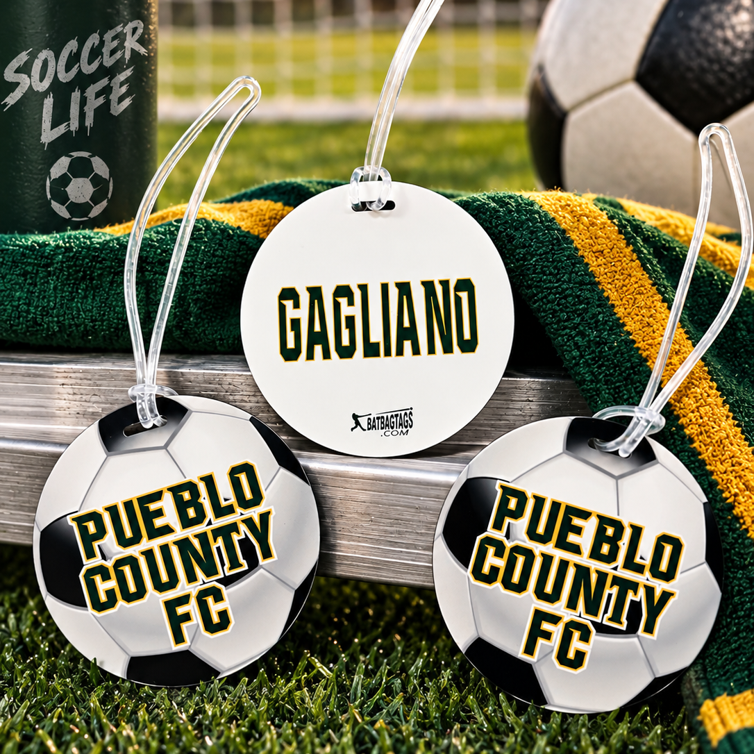 Three round soccer-themed luggage tags displayed on artificial grass with a green and yellow striped towel, a soccer ball, and a water bottle in the background. Two tags are printed as black-and-white soccer balls with yellow 'PUEBLO COUNTY FC' text, and one tag is a white circle with 'GAGLIANO' in yellow.