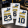 Three white baseball-style towel or flag-style accessories with player names and numbers hang on a locker shelf. The towels show the Titans logo and the names Aston 6 and Turner 10, with matching team decor around them including a helmet, baseball, glove, bat, and a sign reading Titan Pride.