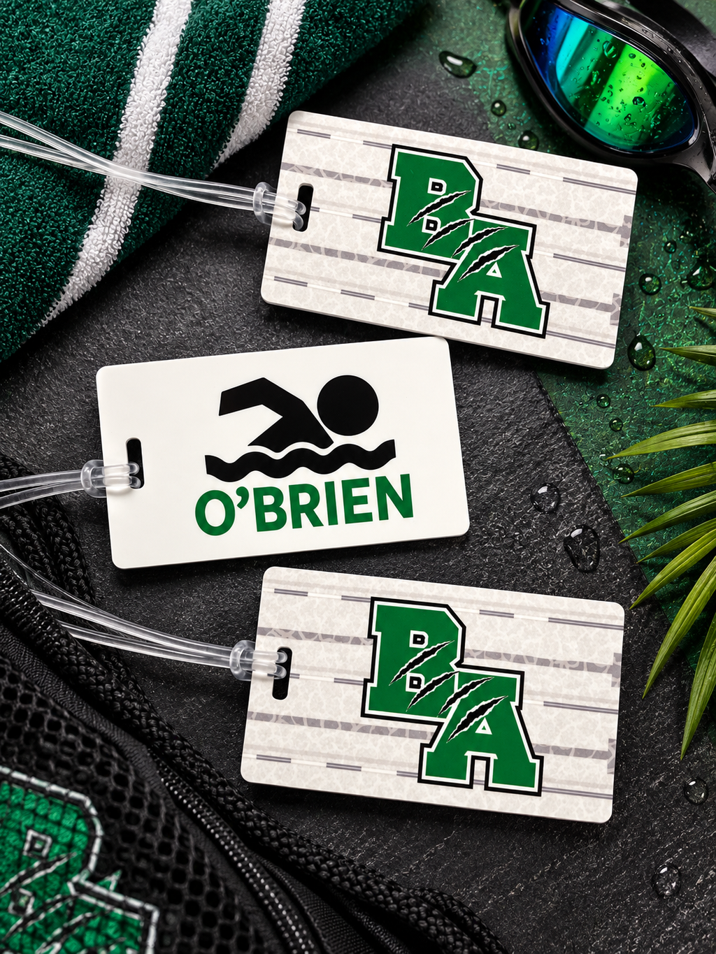 Three custom luggage tags arranged on a dark textured surface with a green and white striped towel, swim goggles, and water droplets. Two tags feature a green and white wood-grain pattern with the letters 'BA' in green, and one tag is solid white with a black swim icon and the name 'O'BRIEN' in green.